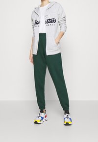 Sudadera gris con cremallera y logo negro, pantalones de chándal verdes y zapatillas deportivas multicolores. Tejido suave, corte relajado y puños elásticos.
