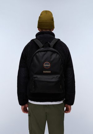 Osoba v zelené čepici, černé fleecové bundě, olivových nákladních kalhotách, s černým batohem Napapijri, otočená zády na jednobarevném pozadí.