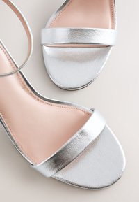 Sandales métalliques argentées à bout ouvert avec brides à la cheville et semelles intérieures couleur nude, vues de dessus sur un fond pâle.