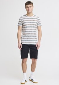 Striped t-shirt in white, green, and brown, paired with black shorts. Model wearing white sneakers with green and black accents.