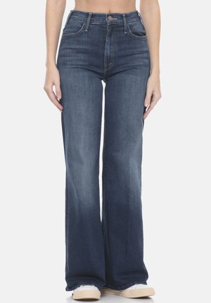 Jeans blu scuro a vita alta e vestibilità slim, leggermente scoloriti, indossati da una persona in piedi con le mani rilassate ai lati, abbinati a sneakers bianche.