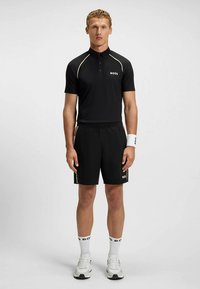 Μαύρο πόλο πουκάμισο με χρυσές λεπτομέρειες, σε συνδυασμό με μαύρο σορτς. Διαθέτει λευκό λογότυπο στο πουκάμισο και στις κάλτσες, μοντέρνο αθλητικό σχέδιο και αθλητικά παπούτσια.