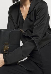 Svart texturerad läderhandväska med guldfärgad hårdvara, utrustad med axelrem och en strukturerad form, placerad bredvid en svart blus med volanger.