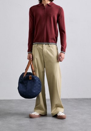 Person iført en vinrød langærmet polo, beige bukser med vide ben, hvide sneakers, der holder en rund navyblå taske med læderstropper.