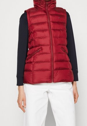 Bezrekawnik pikowana czerwona kamizelka z przednim zamkiem błyskawicznym i dwoma zamykanymi na zamek kieszeniami, noszona na czarnej bluzce z długim rękawem i białych spodniach.