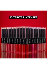Vingt rouges à lèvres dans des tubes transparents avec des bouchons noirs, présentant une variété de teintes allant du nude au rouge vif, disposés en gradient sur un fond rouge.
