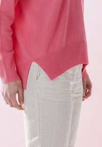 Leichter pinkfarbener Pullover mit gerippten Bündchen und einem gespaltenen Saum, kombiniert mit hellbeigen, strukturierten Hosen. Der Hintergrund ist sanft pink.