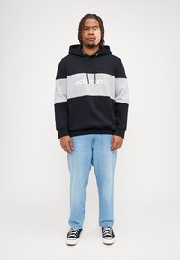 Zwart en grijs kleurgeblokte hoodie met een logo aan de voorkant, een trekkoordkap en een kangoeroezak, gecombineerd met lichtblauwe jeans en zwarte sneakers.