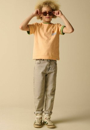 Enfant aux cheveux blonds bouclés portant un T-shirt orange, un jean gris clair, des baskets, ajustant des lunettes de soleil marron, debout devant un fond beige.