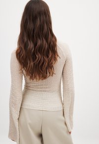 Ilgomis rankovėmis smėlio spalvos marškiniai su tekstūruota bangų raštu ir prigludusia forma, derinami su šviesiomis kelnėmis ir banguotais plaukais fone.