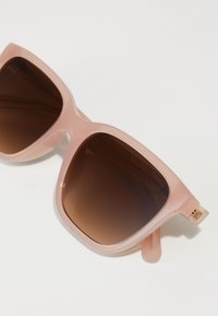 Óculos de sol de plástico rosa com uma grande armação quadrada e lentes castanhas em degradê. Apresenta um detalhe dourado na dobradiça e um acabamento brilhante suave.