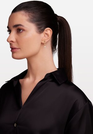 Boucles d'oreilles en or avec un design en barre verticale, présentant un petit détail en perle. Le mannequin porte une chemise en satin noir avec un col.