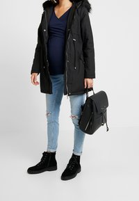 Gravid kvinna i svart parkas, marinblå topp, slitna ljusa jeans, svarta snörstövlar, som håller i en svart läderryggsäck.