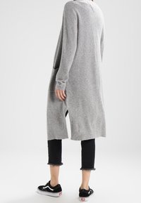 Lång grå stickad cardigan med sidov slit, sänkt axelsöm och fickor. Kombinerad med svarta cropped jeans och svarta sneakers.