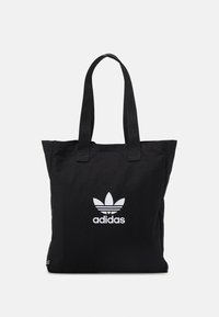 Černá taška přes rameno s dlouhými popruhy, která má uprostřed na přední části bílou trefoil logo Adidas na čistém pozadí.