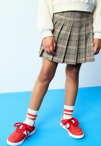 Karierte Plissee-Rock in Beige und Braun, kombiniert mit roten Wildledersneakern und weißen Socken mit roten Streifen. Glatte Oberfläche und gestuftes Design.