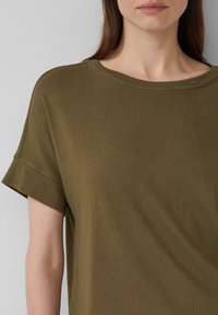 Olive green, krótki rękaw, luźna koszulka z okrągłym dekoltem. Miękka tekstura i widoczne szwy na podwiniętych rękawach.