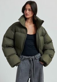 Puffig olivgrön jacka med hög krage, dragkedja och ribbade ärmar. Buren över en svart topp och matchad med grå högmidjade byxor.