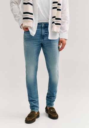 Mężczyzna ubrany w jasnoniebieskie jeansy typu slim fit, białą koszulę, pasiasty sweter zarzucony na ramiona i brązowe mokasyny, stojący na jednolitym tle.