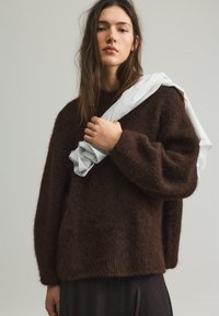 Bruine, pluizige trui van zacht materiaal, met losse mouwen en een ronde halslijn. Over één arm is een wit gestreepte stof gedrapeerd.