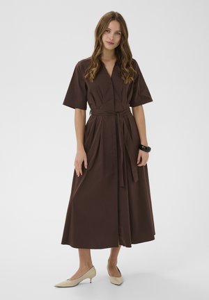 Kobieta z długimi falowanymi włosami w brązowej sukience midi z paskiem, beżowych butach na niskim obcasie i czarnej bransoletce, stojąca na jednolitym tle.