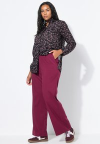 Zwarte shirt met knoopsluiting en een roze bloemenprint, gecombineerd met losse bordeauxrode broeken. Sneakers met witte accenten maken de outfit af.