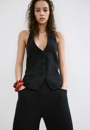 Jeune femme aux cheveux bouclés portant un top noir sans manches boutonné et un pantalon noir, mains dans les poches, et un gros bracelet rouge à perles au poignet gauche.