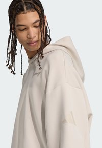 Felpa color crema con grande cappuccio, texture morbida, vestibilità rilassata e un discreto dettaglio logo a tre righe sulla manica sinistra.