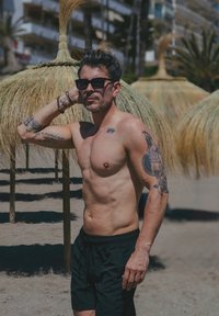 Mężczyzna bez koszulki z tatuażami, ubrany w czarne kąpielówki, stoi na plaży obok słomianych parasoli, z palmami i budynkami w tle.