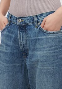 Personne portant un jean en denim bleu ample avec les mains dans les poches avant et un haut côtelé marron clair rentré.