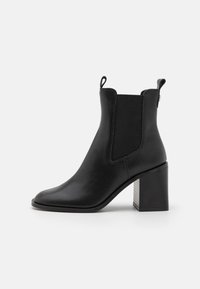 Bottine en cuir noir avec panneaux latéraux élastiques, languettes de tirage, bout pointu et talon bloc épais sur fond blanc.