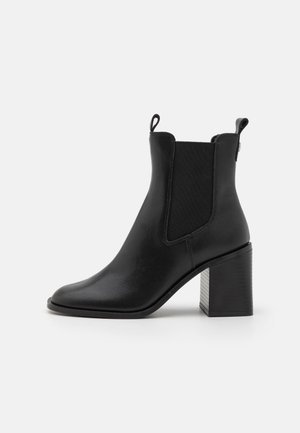 Bota de tobillo de cuero negro con paneles elásticos en los laterales, lengüetas para tirar, punta puntiaguda y tacón bloque grueso sobre un fondo blanco.