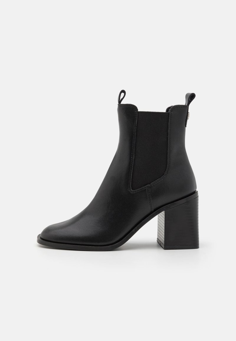 Bottine en cuir noir avec panneaux latéraux élastiques, languettes de tirage, bout pointu et talon bloc épais sur fond blanc.