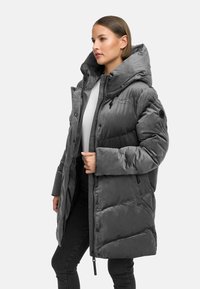 Sieviete, tērpusies vidēja garuma pelēkā pufīgā mētelī ar lielu kapuci, apģērbu apvienojusi ar melnām biksēm un baltu topu, stāvot pret vienkāršu fona.