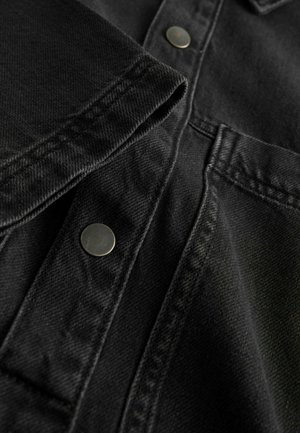 Melnas denima krekla tuvplāns, kurā redzama teksturēta auduma virsma, redzami šuvju elementi un sudraba metāla pogas gar iešu un apkakli.