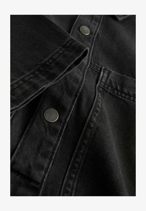 Melnas denima krekla tuvplāns, kurā redzama teksturēta auduma virsma, redzami šuvju elementi un sudraba metāla pogas gar iešu un apkakli.