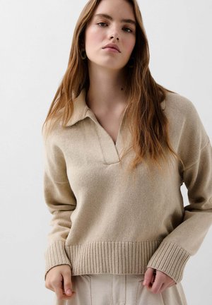 Beige Strickpullover mit Kragen, gerippten Bündchen und Saum. Weiche Textur und lässige Passform. Das Model steht vor einem neutralen Hintergrund.