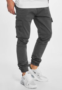 Man in donkergrijze cargo broek en witte Nike Air Max 270 sneakers, staand tegen een effen lichte achtergrond.