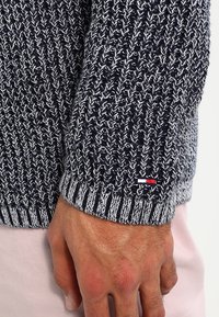 Marineblauer und weißer gestrickter Pullover mit strukturiertem Muster, gerippten Bündchen und einem kleinen Logo-Patch am Handgelenk.