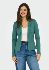 Blazer teal avec une coupe ajustée, fermeture à un bouton et deux poches frontales. Assorti à un top blanc et un jean bleu taille haute.