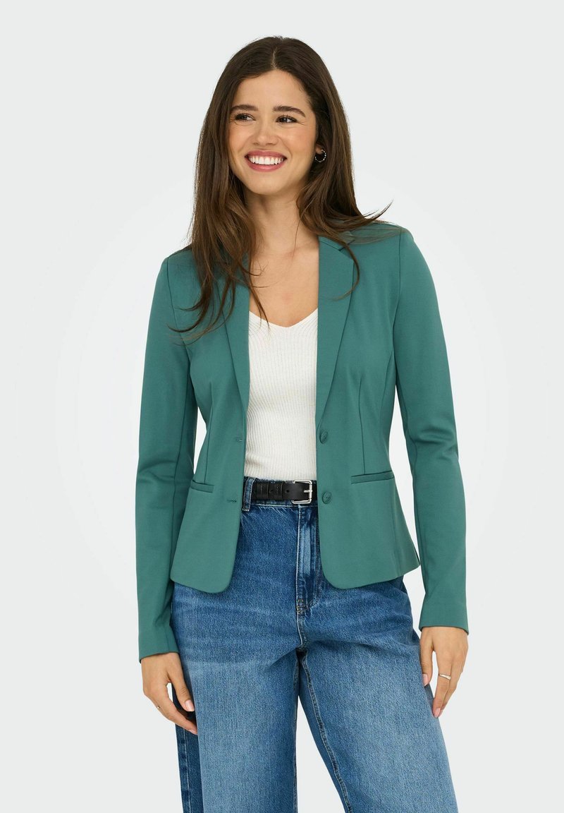 Blazer teal avec une coupe ajustée, fermeture à un bouton et deux poches frontales. Assorti à un top blanc et un jean bleu taille haute.
