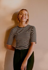 Rövid szőke hajú nő fekete-fehér csíkos ingben, mosolyogva áll egy bézs fal előtt, mögötte árnyék vetül.