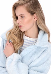 Jachetă din fleece albastru deschis cu guler înalt, textură moale și închidere cu fermoar. Bluză cu dungi dedesubt, model care ține jacheta.