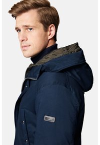 Granatowa kurtka z kapturem z pikowaną zieloną podszewką, srebrnymi akcesoriami i naszywką z logo na lewym rękawie. Gładka faktura z dopasowanym krojem.