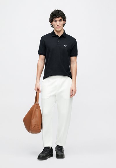 Čierne polokošeľa s jemným logom, v kombinácii s bielymi zúženými nohavicami. Muž drží hnedú koženú tašku a má na sebe čierne tenisky.