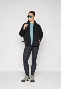 Vrouw in zwarte fleece jack, teal shirt, zwarte leggings, grijze wandelschoenen, blauw-getinte zonnebril, die een zwarte rugzak draagt, staand tegen een witte achtergrond.