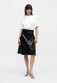 Γυναίκα που φοράει λευκό πουκάμισο με φουσκωτά μανίκια, μαύρη κομψή υφασμάτινη φούστα και ασημένια μυτερά παπούτσια mule, στέκεται μπροστά σε απλό ανοιχτό φόντο.