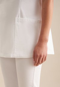 Tunika bianca con orlo curvo e tasca laterale, realizzata in un tessuto liscio. Accompagnata da pantaloni bianchi attillati, che evidenziano un design pulito.