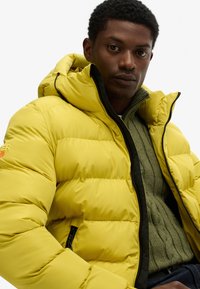 Geel puffer jack met een capuchon, voorzien van een ritssluiting, horizontaal gestikt patroon en zwarte accenten, gedragen over een groene gebreide sweater.
