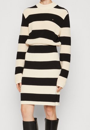 Kobieta ubrana w czarny i beżowy sweter z szerokimi poziomymi paskami oraz pasującą prążkowaną spódnicę, do tego czarne kozaki za kolano.